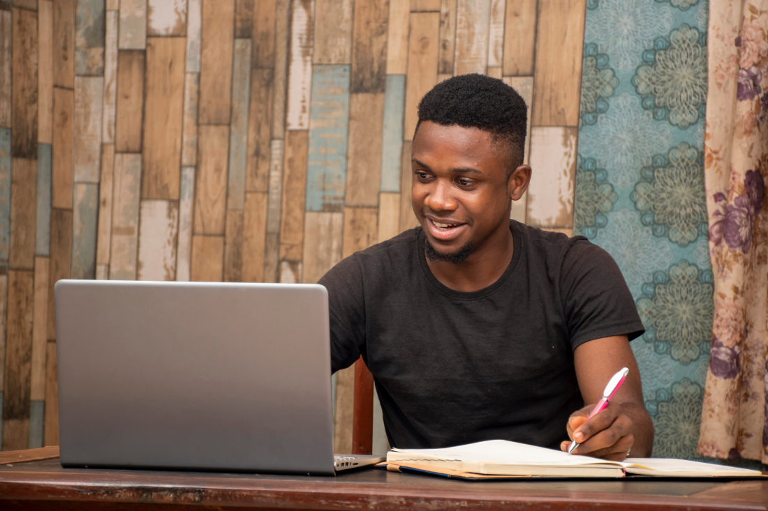young african man studying at home using his laptop, receiving lectures online and taking notes, smiling